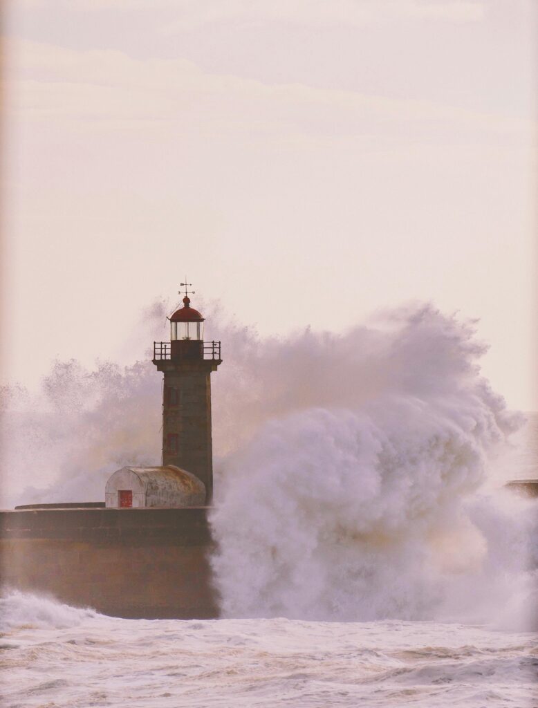 Powerful ocean waves crash against a solitary lighthouse at sunset, creating a dramatic and captivating scene.